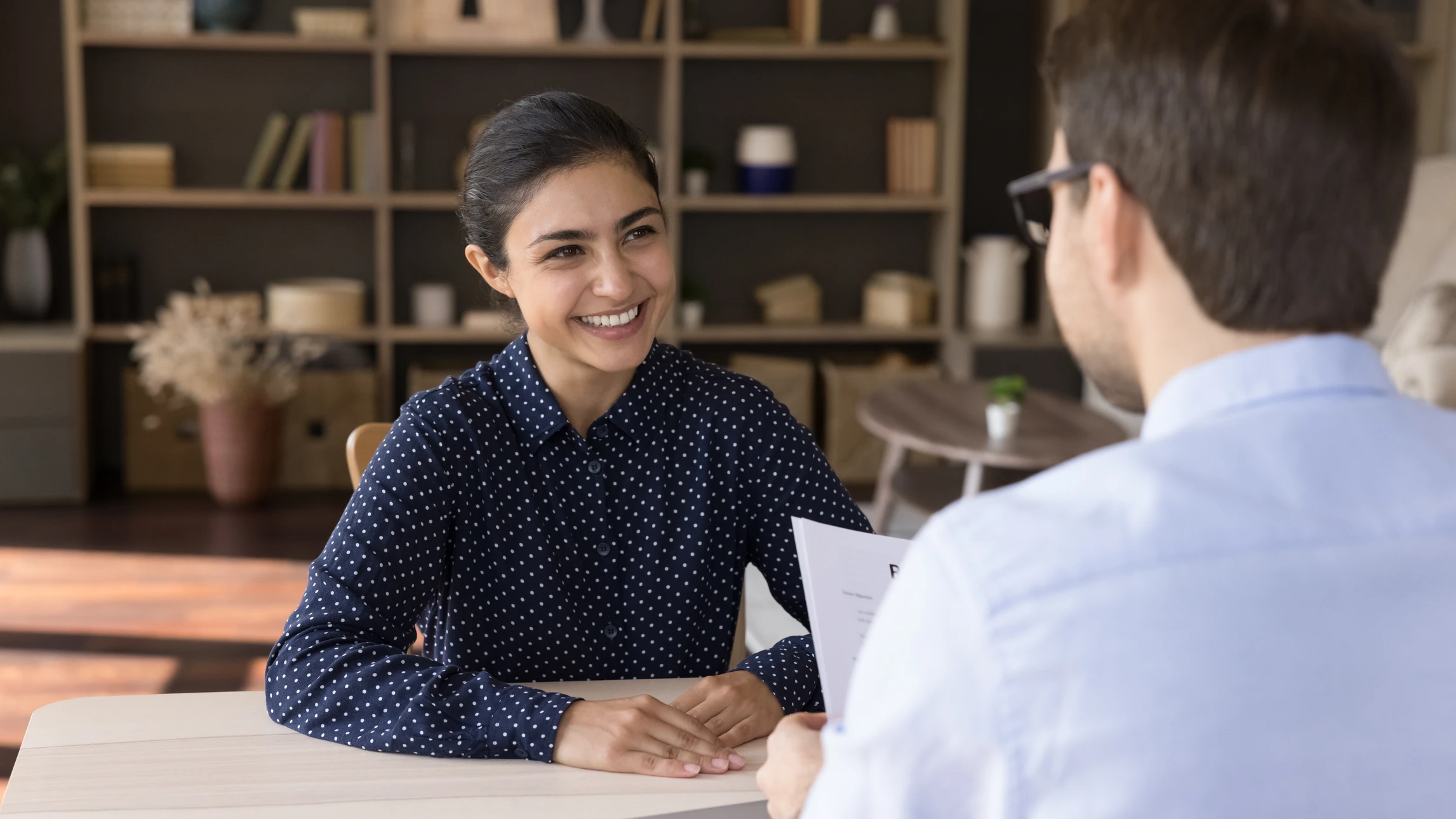 Femme souriante en chemisier bleu à pois blancs assise face à un homme en chemise claire lors d'un entretien professionnel dans un bureau moderne avec étagères en arrière-plan