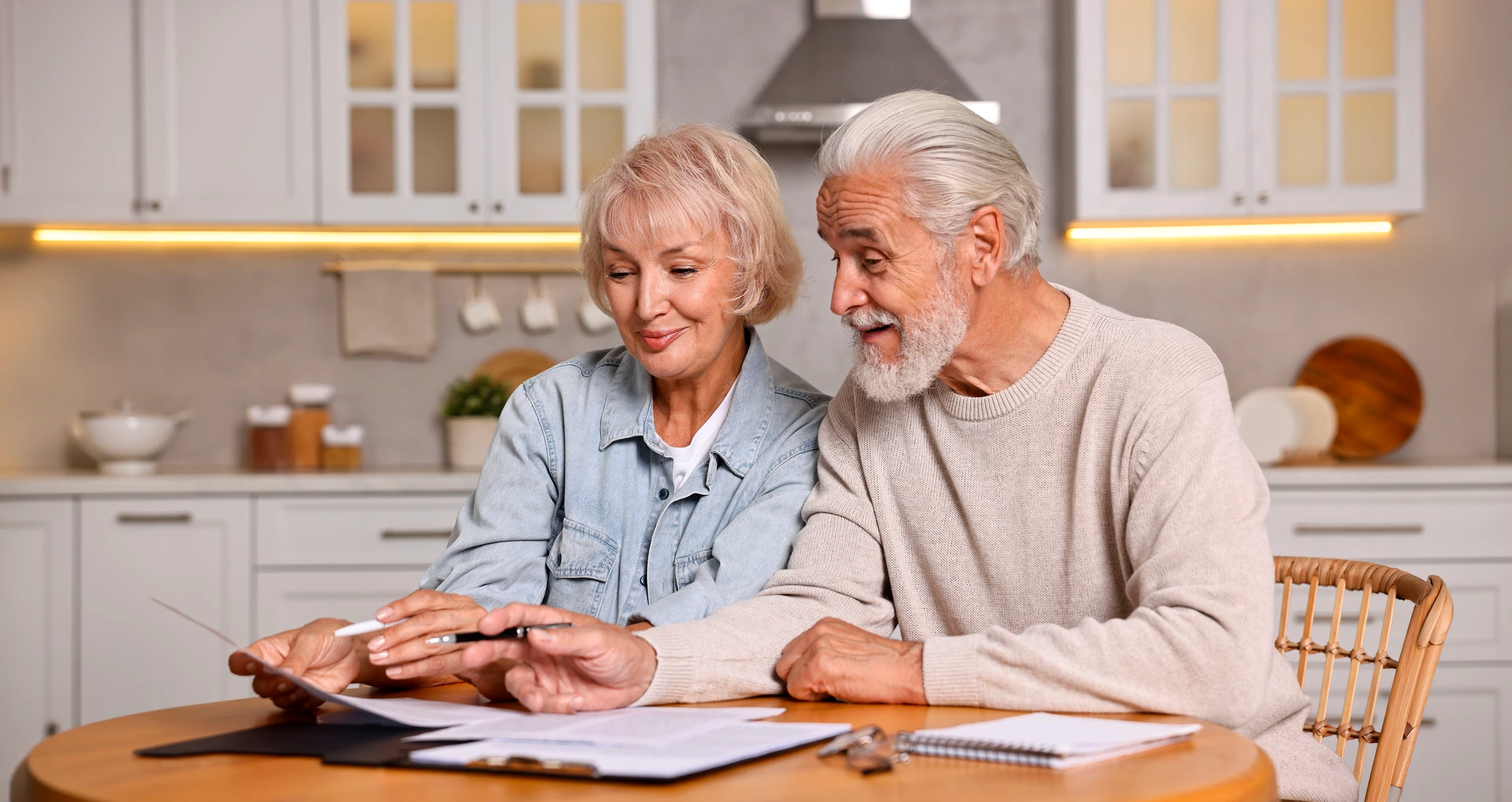 Photographie d'un conseiller en assurance d'âge mûr, vêtu professionnellement, assis à son bureau dans un cadre de bureau moderne et accueillant. Il consulte des documents financiers ou un dossier avec un client senior assis face à lui. La scène capture un moment de conseil bienveillant et confiant, mettant l'accent sur la relation de confiance et la planification sereine de l'avenir. L'éclairage naturel traversant de grandes fenêtres crée une atmosphère rassurante et optimiste.