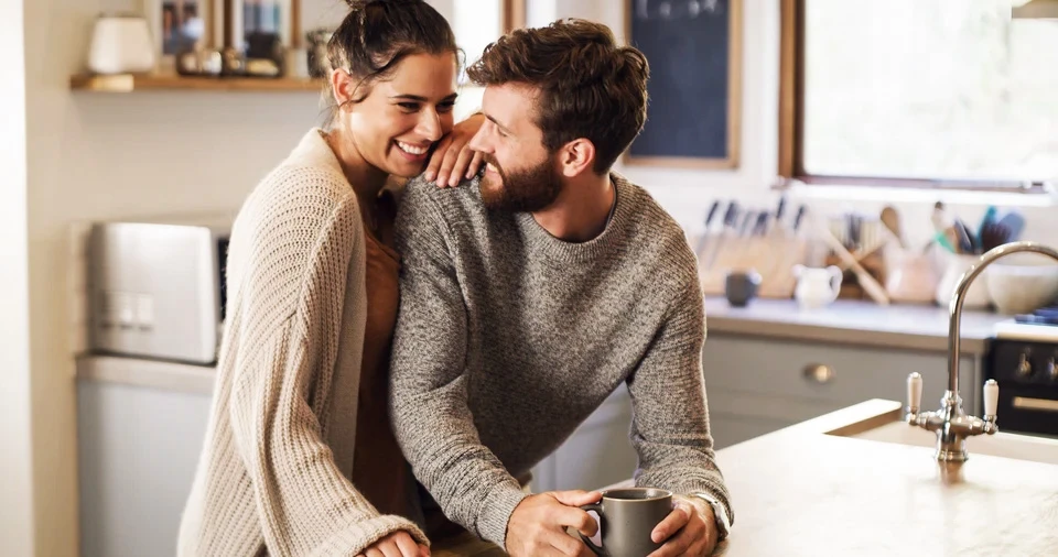 Couple mature souriant dans un intérieur lumineux, femme blonde enlace homme aux cheveux grisonnants, tous deux vêtus de vêtements décontractés clairs, expression de bonheur et complicité