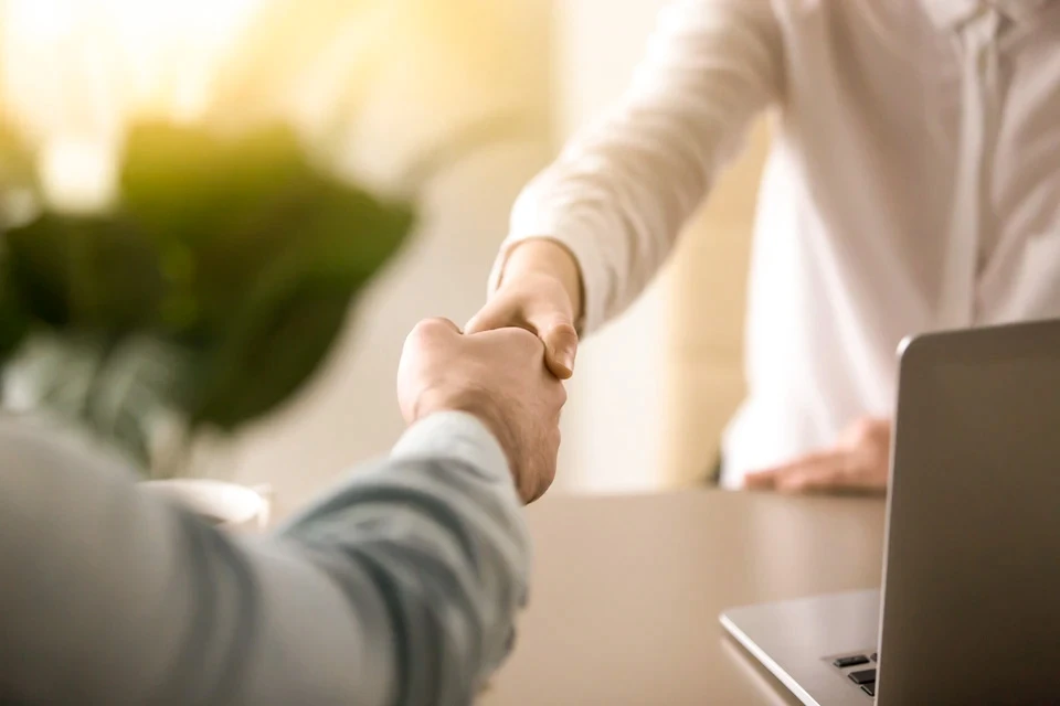 Deux personnes se serrent la main dans un bureau moderne, une en chemise blanche debout, l'autre en pull gris assis