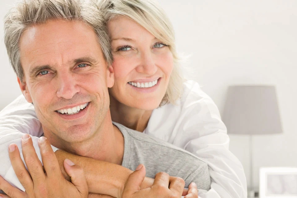 Couple mature souriant dans un int&eacute;rieur lumineux, femme blonde enlace homme aux cheveux grisonnants, tous deux v&ecirc;tus de v&ecirc;tements d&eacute;contract&eacute;s clairs, expression de bonheur et complicit&eacute;