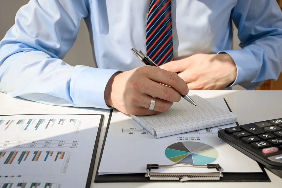 Homme d'affaires mature en costume trois-pièces gris à carreaux, barbe grise soignée, tenant des lunettes, posant avec assurance dans un bureau moderne aux baies vitrées